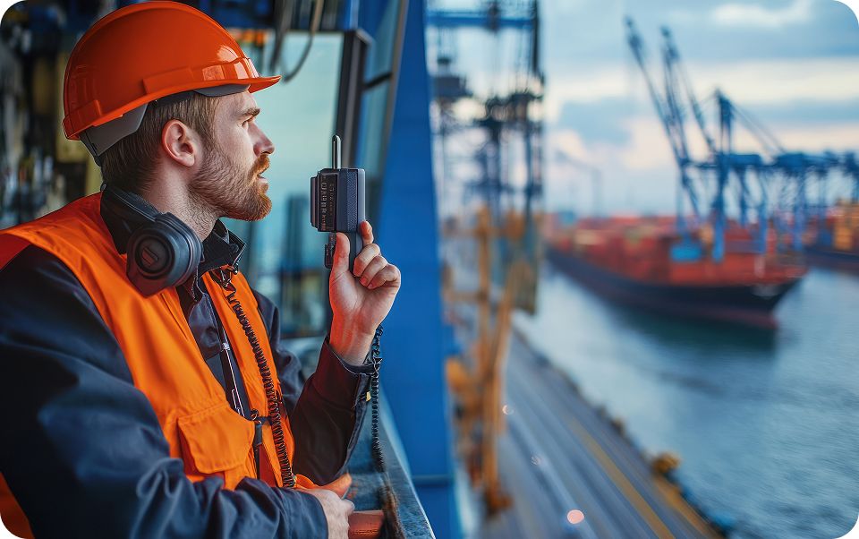 A port worker in an orange safety vest and helmet with a radio monitors a ship in the port